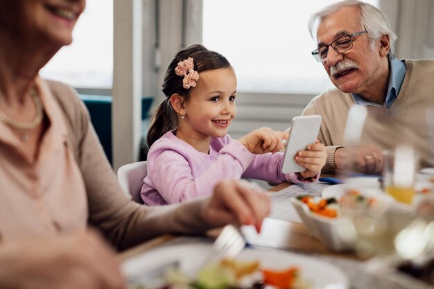 Happy little girl having lunch with her grandparents and using smart phone at dining table