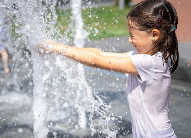 Happy little girl among the splashing water of the city fountain has fun and escapes from the heat.