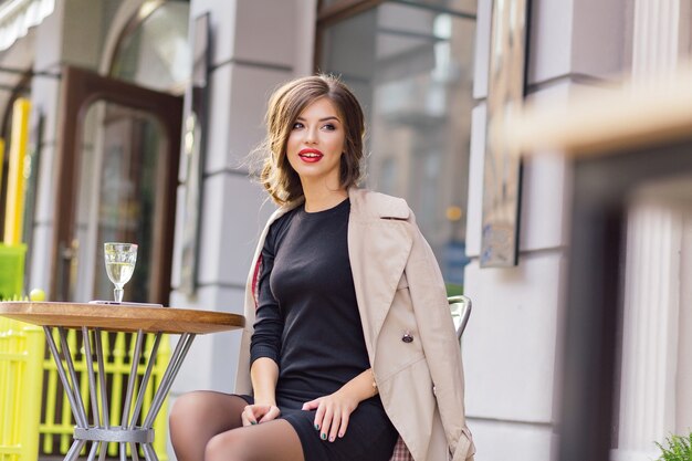 Happy lovable woman in black dress and beige coat sitting in outdoor cafeteria and resting with a glass of wine