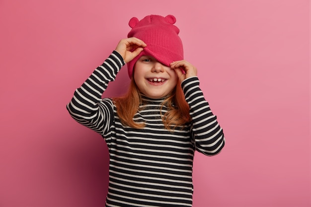 Happy lovely girl preschooler looks from under hat, plays hide and seek, wears casual striped jumper, isolated over rosy pastel wall, has healthy skin, hears hilarious story, laughs positively