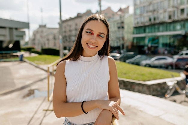 Happy lovely girl with long hair and bright make up is wearing white tshirt smiling at camera and spend time outdoor in sunny warm day