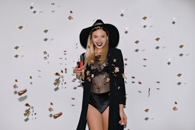 Happy lovely woman with blond hair posing with wine over isolated wall with confetti