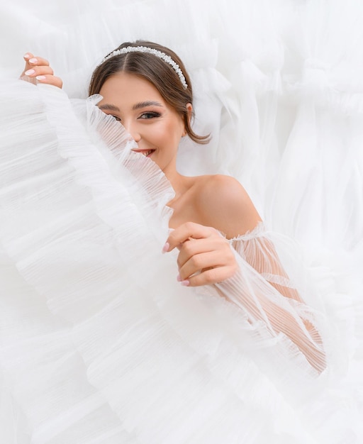 Happy lovely young woman lying on her wedding white puffy dress looking into camera Smiling bride girl with perfect nude makeup and jewelry Wedding dress concept Adorable bride Wedding salon