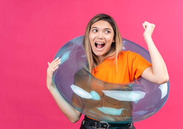 Free Photo a happy lovely young woman in an orange t-shirt raising clenched fist while standing on inflatable ring on a pink wall