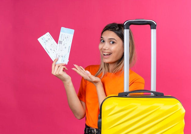 Free photo a happy lovely young woman in an orange t-shirt showing plane tickets with yellow suitcase on a pink wall