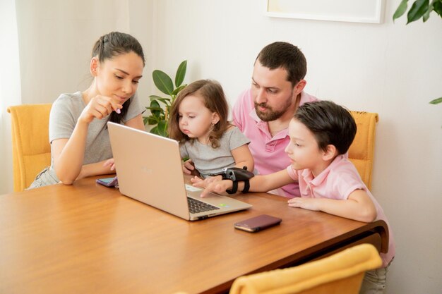 Happy loving family couple parents and little cute children son and daughter sitting at the table together father working on laptop with busy face working at home concept