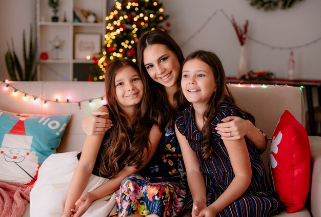 happy loving two sisters and young mother at home at christmas time sitting on sofa in living room mother hugging daughters elder daughter looking at side younger one and mother looking at camera