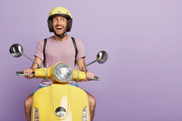 Free Photo happy male biker or courier drives yellow scooter, wears protective helmet, casual t shirt, poses on his own transport, looks joyfully aside, transports something, isolated on purple wall, blank space