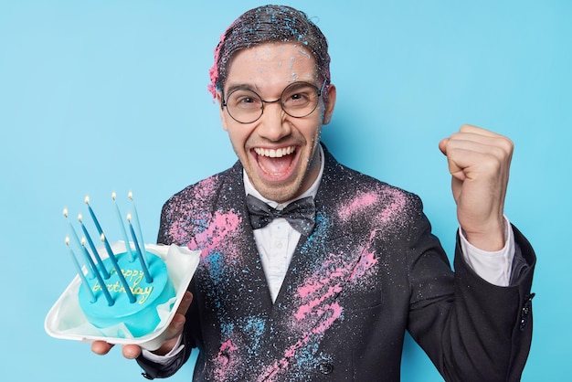 Happy man celebrates birthday with friends on arty holds festive cake clenches fist with triumph wears formal suit expresses positive emotions isolated over blue background People and celebration