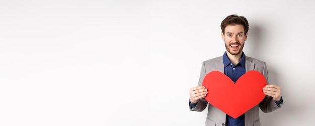 Free photo happy man confessing in love showing heart cutout and smiling at camera standing in suit on romantic