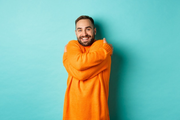 Happy man feeling comfortable, wearing warm sweater and hugging himself, smiling satisfied, standing over light turquoise wall.