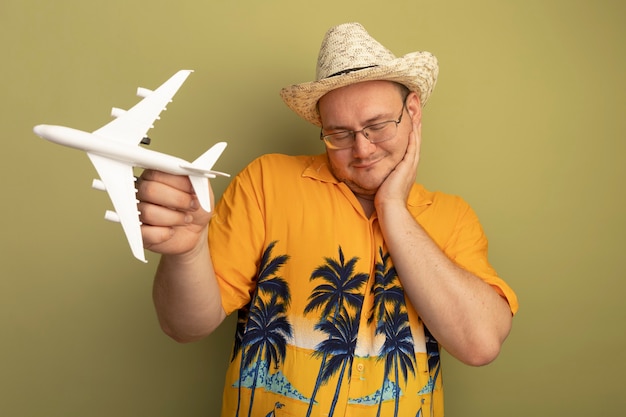 Free Photo happy man in glasses wearing orange shirt in summer hat holding toy airplane smiling with hand on his cheek standing over green wall