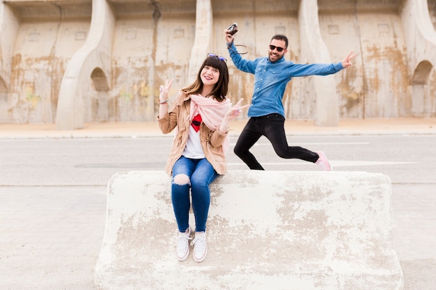 Happy man holding camera jumping in front of woman sitting over bench