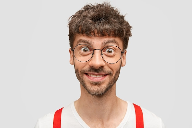 Free photo happy man looks with cheerful and intriguing expression, rejoices his success, wears round spectacles and white clothes with red braces