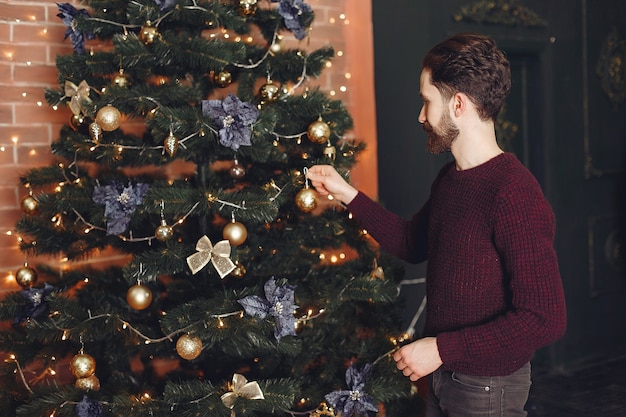 Happy man in red sweater. Guy in front of the fireplace. Male on the background of Christmas tree.