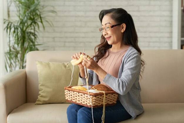 Happy Mature Asian Woman Knitting Clothes Sitting On Sofa At Home