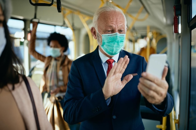 Happy mature businessman with face mask having video call over mobile phone in a bus