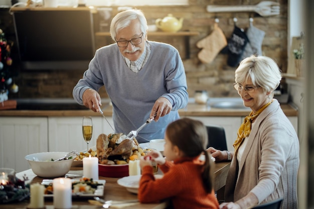 Happy mature man carving roasted Thanksgiving turkey at dining table