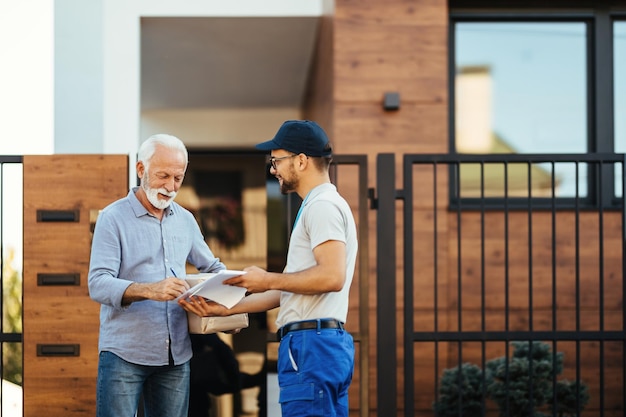 Free Photo happy mature man receiving package from a courier and signing paperwork in front of his house