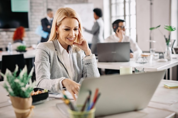 Happy mid adult businesswoman using computer and surfing on the Internet while working in the office There are people in the background