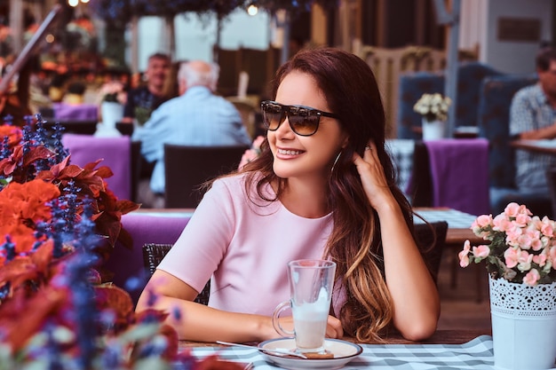 Free Photo happy middle age businesswoman with long brown hair wearing a pink dress sitting with a glass of cappuccino at the outdoor cafe.