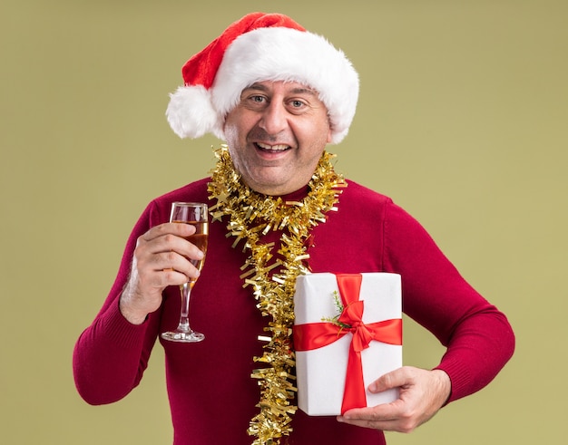 Happy middle age man wearing christmas santa hat with tinsel around neck holding christmas  present and glass of champagne looking at camera with smile on face  standing over green  background