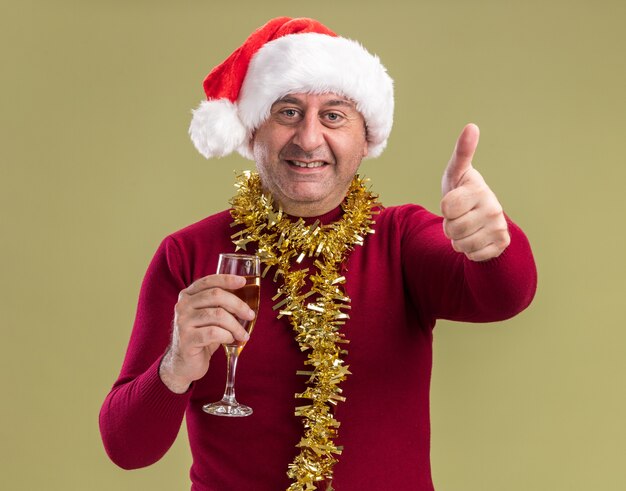 Happy middle age man wearing christmas santa hat with tinsel around neck holding glass of champagne looking at camera smiling showing thumbs up  standing over green  background