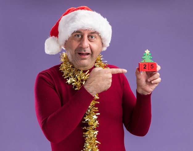 Happy middle age man wearing christmas santa hat  with tinsel around neck holding toy cubes with date twenty five pointing with index finger at them smiling standing over purple background