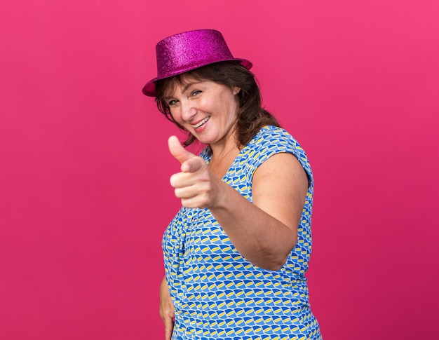 Happy middle age woman in party hat pointing with index finger  smiling cheerfully celebrating birthday party standing over pink wall