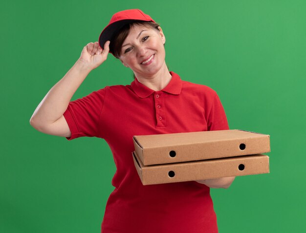 Happy middle aged delivery woman in red uniform and cap holding pizza boxes looking at front smiling confident standing over green wall