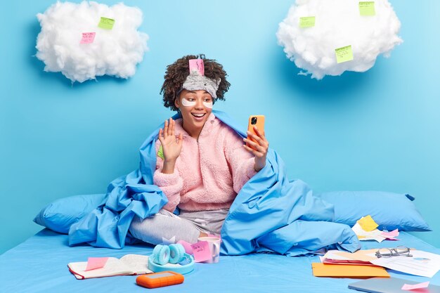 Happy millennial girl with Afro hair waves hello in smartphone device does reserach or assignment at home enjoys domestic atmosphere