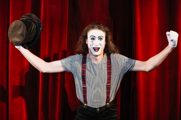 Happy mime standing in front of red curtain holding hat flexing his muscle