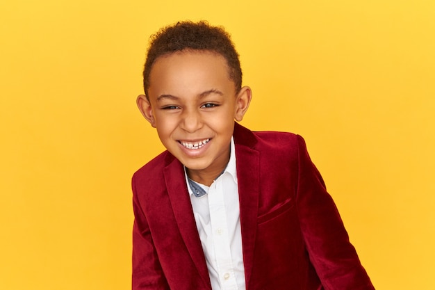 Free photo happy mischievous adorable black african boy in stylish velvet jacket being in good mood, laughing at funny story, joke or prank