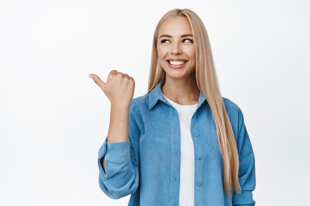 Happy modern girl pointing and looking left with satisfied smile recommending shop showing store advertisement sale banner standing over white background