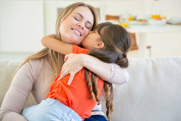 Happy mom sitting with her little girl on couch, holding kid in arms and hugging her.