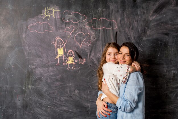 Happy mother and daughter hugging near chalkboard with drawing 