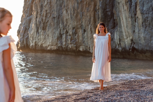 Happy mother and daughter in white dress standing face to face in seashore during sunset .