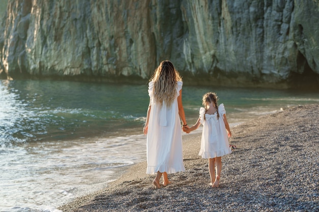 Happy mother and daughter in white dress walking in seashore during sunset. back view