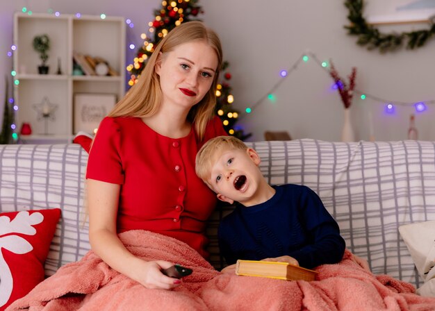 Happy mother in red dress with her little child under blanket reading book in a decorated room with christmas tree in the wall