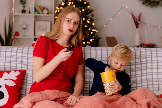 Happy mother in red dress with her little child sitting on a couch under blanket with bucket of popcorn watching tv together in a decorated room with christmas tree in the wall