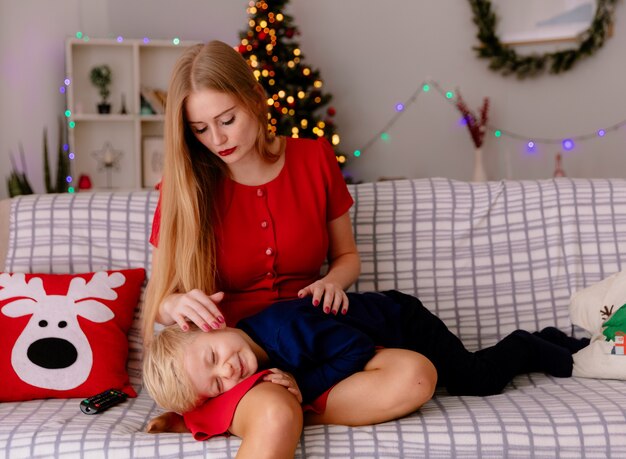 happy mother in red dress with her little child who laying on her knees on a couch having fun watching tv together in a decorated room with christmas tree in the background