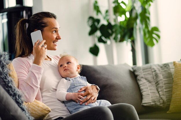 Happy mother talking on the phone while holding her baby and relaxing on sofa in the living room