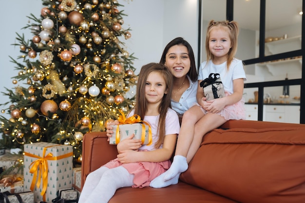 Happy mother and two little daughters are sitting on sofa and smiling against background of Christmas tree