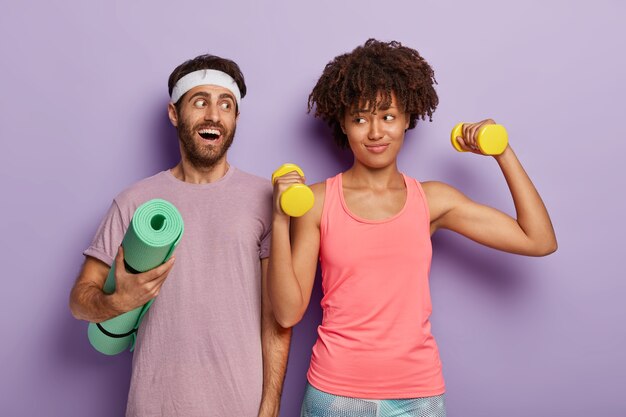 Happy multiethnic couple achieve sport success, have workout in gym with dumbbells