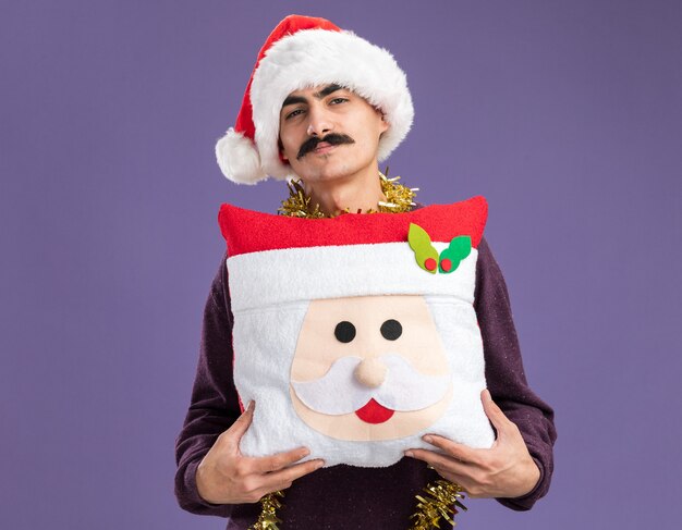 Happy mustachioed man wearing christmas santa hat  with tinsel around his neck holding christmas pillow  with smile on face standing over purple wall