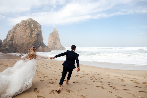 Happy newlyweds holding for their hands are running across the beach on the Atlantic ocean