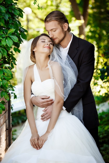 Happy newlyweds smiling with closed eyes, embracing in park.