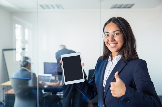 Happy office lady showing blank tablet screen, making like gesture, looking at camera and smiling. Copy space. Communication and advertising concept