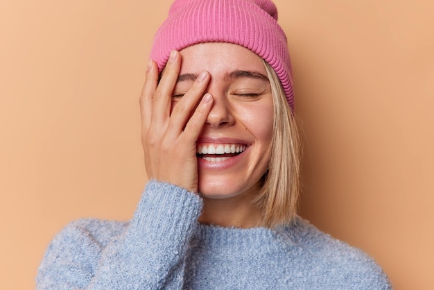 Free photo happy optimistic young woman makes face palm smiles broadly keeps eyes closed being in good mood wears hat and sweater isolated over beige background positive emotions and feelings concept