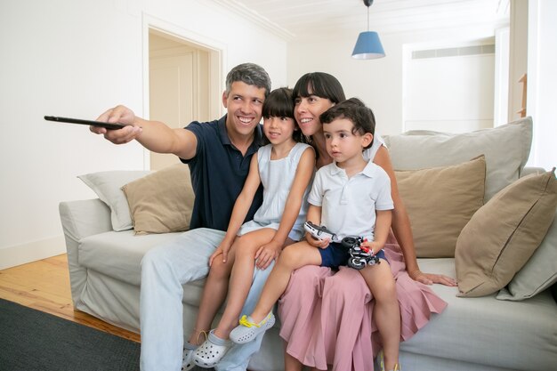 Happy parent couple with two kids watching TV, sitting on couch in living room and using remote control.